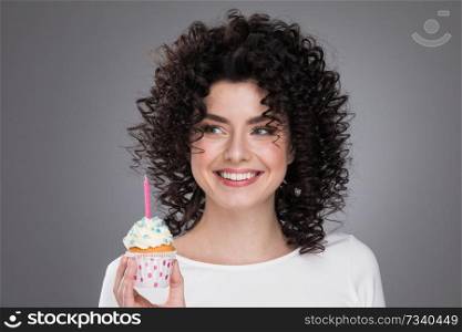 Cheerful beautiful curly young woman making a wish holding birthday cupcake with candle. Woman with birthday cupcake