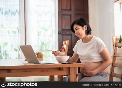 Cheerful Beautiful Pregnant woman enjoy with laptop at table. Happy Young Asian Mother smiling making video call online during having breakfast in dining room at home. Internet communication