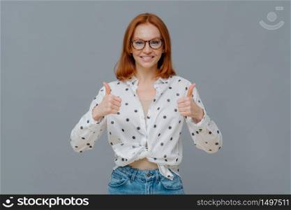 Cheerful beautiful young redhead woman shows thumbs up, likes amazing idea, gives positive feedback about something, smiles positively, wears optical glasses and stylish clothes, gestures indoor
