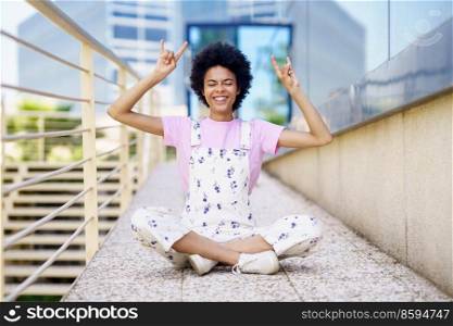 Cheerful black woman in casual clothes woman making the sign of horns with her hands. Happy young femeale, afro hair, in urban background.. Cheerful black woman in casual clothes woman making the sign of horns with her hands.