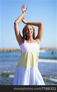 Cheerful black woman in stylish dress raising arms and looking at camera with smile while dancing on blurred background of waving sea and blue sky. Happy African American female dancing near waving sea