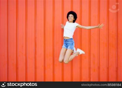 Cheerful black woman looking at camera, while jumping with raised arms against a red urban wall. Full body photograph. Black girl jumping with raised arms against a red urban wall.