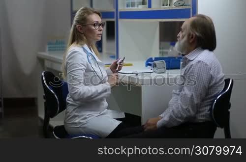 Cheerful blonde female neurologist in eyeglasses and lab coat inspecting senior male patient&acute;s nervous system using the test hammer in hospital. Attractive female physician testing reflexes of elderly man&acute;s eyes with neurological hammer in clinic.