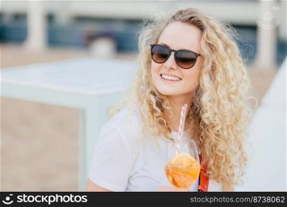 Cheerful blonde female with wavy light hair, positive smile, wears shades, looks aside with happy expression, drinks fresh cocktail, poses against blurred background with copy space for advertisement