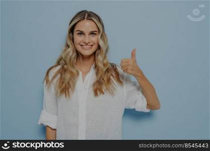 Cheerful blonde girl in white shirt showing thumbs up gesture with her hand while smiling, admitting that she liked that, standing alone next to blue wall. Positive feedback concept. Cheerful blonde girl showing thumbs up gesture