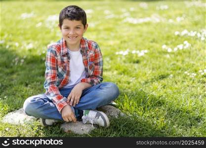 Cheerful boy portrait sitting on city park grass while looking at camera