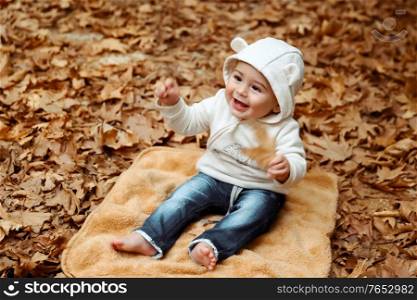 Cheerful boy with pleasure spending time in the autumn park, little baby sitting barefoot on the blanket on the ground covered with dry tree leaves, happy autumn time