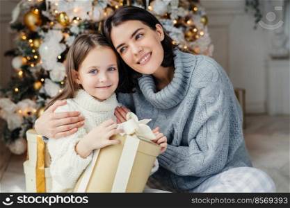 Cheerful brunette female leans at her daugter, embraces her, presents gift box, being in living room near decorated New Year tree. Glad family  mother and daughter in warm sweaters celebrate Christmas