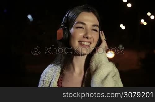 Cheerful brunette girl in headphones singing along with the song, holding earcup with hand while enjoying music in night city. Emotional charming woman listening music with earphones as she spends time outdoors at night. Slow motion. Stabilized shot.