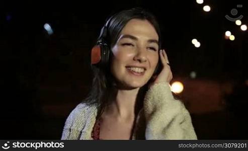 Cheerful brunette girl in headphones singing along with the song, holding earcup with hand while enjoying music in night city. Emotional charming woman listening music with earphones as she spends time outdoors at night. Slow motion. Stabilized shot.