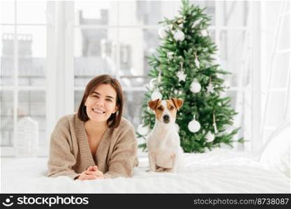 Cheerful brunette woman gets jack russell terrier dog as Christmas gift, lies on bed, feels relaxed and happy, green decorated New Year tree in background. People, animals, holidays concept.