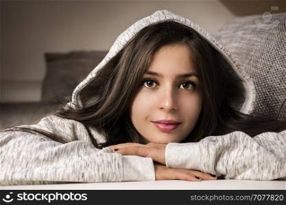 Cheerful brunette woman in a cozy ambiance - Beautiful, young brunette woman, relaxing on the couch, at home, dressed in a cozy hooded sweatshirt, smiling at the camera.