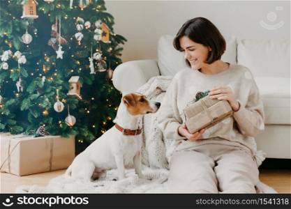 Cheerful brunette woman shows received presents to favourite dog, pose together on floor in cozy room, have festive mood, prepare for Christmas or New Year, enjoy winter time. Hoilday concept