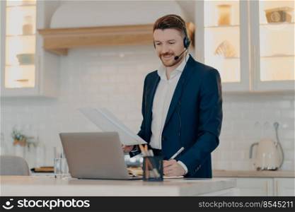 Cheerful business professional in suit holding sheets of paper, reading information and making some notes during online meeting, using laptop and headset while working at home. Remote work concept. Happy executive manager holding document with report while working remotely