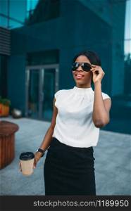 Cheerful business woman in sunglasses with cardboard coffee cup resting during the break outdoors, office building on background. Black businesswoman in skirt and white blouse. Business woman in sunglasses with coffee resting