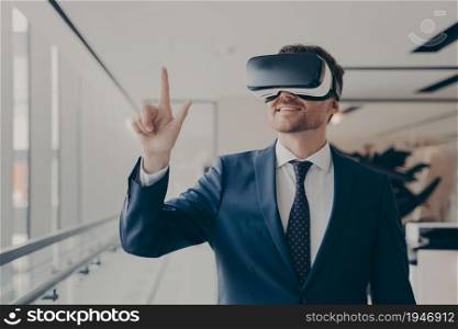 Cheerful businessman dressed formally using vr headset at work, wearing virtual reality glasses and working in augmented reality, pointing at virtual objects with finger. Futuristic technology in business. Cheerful businessman dressed formally using vr headset at work