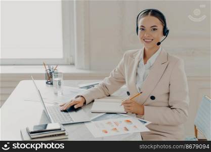 Cheerful businesswoman in formal suit wearing headset with microphone, taking notes and smiling at camera while sitting at work desk with laptop in call center office and working with financial report. Smiling female economist having web conference in office