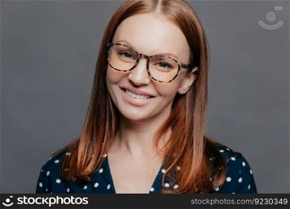 Cheerful close up shot of pretty woman with brown hair, toothy smile, wears spectacles, dressed in formal blouse, poses over grey background, being pleased to meet with colleagues on meeting