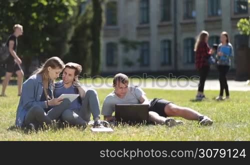 Cheerful college students sitting on green grass in university campus using laptop and tablet while studying outdoors. Attractive student couple working with digital tablet and talking while classmate typing on laptop as they stydy on park lawn.