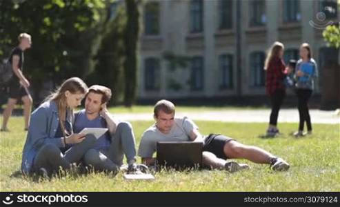 Cheerful college students sitting on green grass in university campus using laptop and tablet while studying outdoors. Attractive student couple working with digital tablet and talking while classmate typing on laptop as they stydy on park lawn.