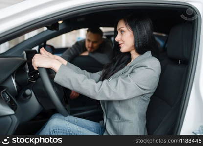 Cheerful couple buying new car in showroom, woman behind the wheel. Male and female customers choosing vehicle in dealership, automobile sale, auto purchase