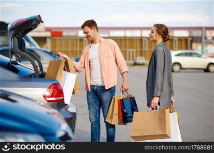Cheerful couple puts their purchases in the trunk on supermarket car parking. Happy customers carrying purchases from the shopping center, vehicles on background. Couple puts their purchases in trunk on parking