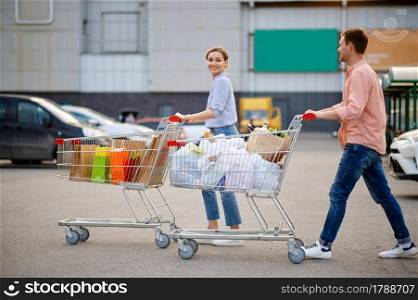 Cheerful couple with bags in carts on supermarket car parking. Happy customers carrying purchases from the shopping center, vehicles on background. Cheerful couple with bags in carts on parking