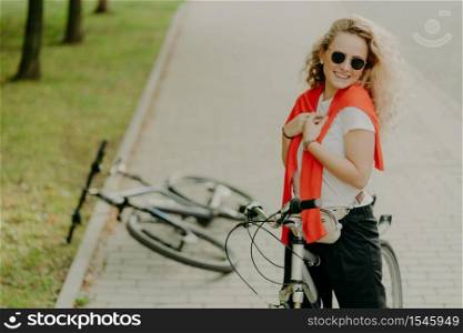 Cheerful curly haired female stops during way, poses on bicycle at road, friends bike lies near, wears sunglasses, t shirt and trousers, carries little bag around waist, being fit and healthy