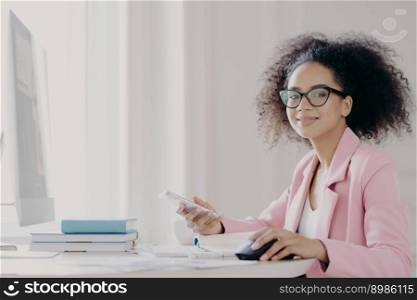 Cheerful dark skinned lady holds mobile phone in hand, waits for call, sits in front of computer, wears optical glasses and elegant suit, poses in her cabinet being busy at work. Technolgy, occupation