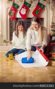 Cheerful daughter and mother wrapping sweater in wrapping paper at Christmas eve