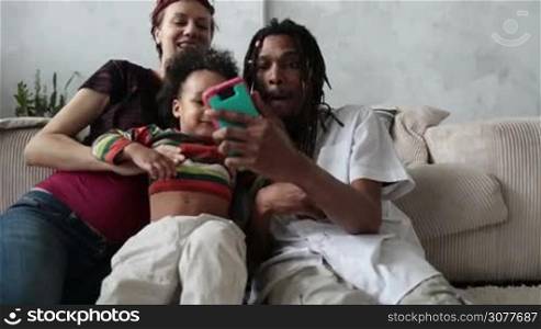 Cheerful diverse family with mixed race curly toddler son making self portrait with mobile phone and smiling while sitting together on the floor in modern apartment. Slow motion.