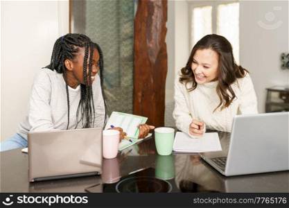 Cheerful diverse female freelancers sitting at table with documents and laptops while working on startup project together. Multiracial women working together from home