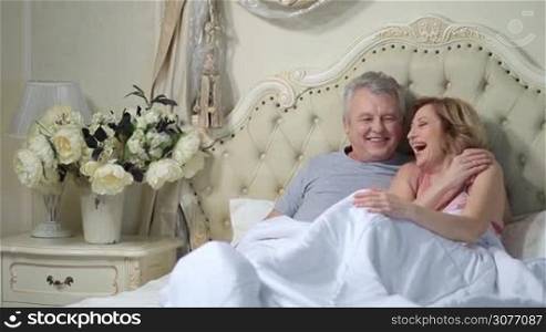 Cheerful elderly couple sitting on bed at home and laughing. Happy mature loving couple lounging in the bedroom after awaking. Senior man telling funny jokes to his charming wife and laughing.