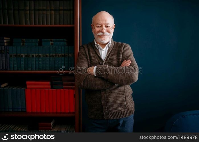 Cheerful elderly man in glasses poses at bookcase in home office. Bearded mature senior poses in living room, old age businessman. Cheerful elderly man in glasses poses at bookcase