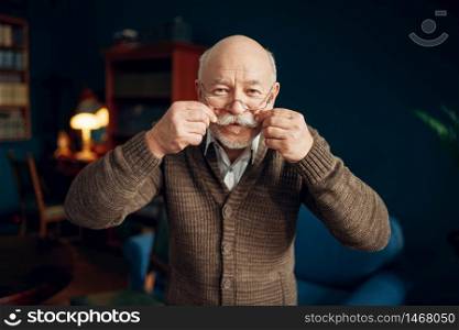 Cheerful elderly man shows his moustache in home office. Bearded mature senior poses in living room, old age businessman. Cheerful elderly man shows his moustach