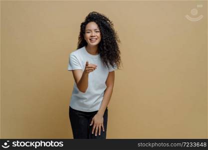 Cheerful energetic young woman with curly Afro hair points index finger forward at camera, feels positive and chooses someone, wears white t shirt and jeans, notices something nice in front.