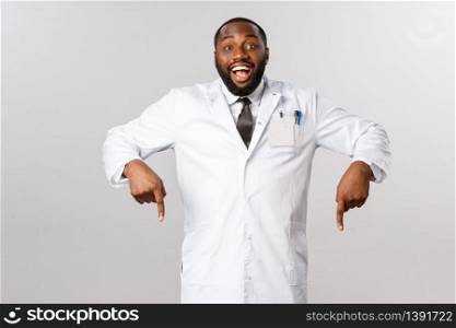 Cheerful excited, happy african-american male doctor inviting see great thing, pointing fingers down, ask to look and check-out awesome product, guarantee best quality, grey background.. Cheerful excited, happy african-american male doctor inviting see great thing, pointing fingers down, ask to look and check-out awesome product, guarantee best quality, grey background