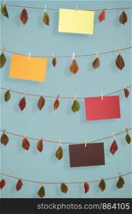 Cheerful fall background with autumn leaves and different colored empty paper sheets, hanging on strings with wooden clips, on a blue wall.
