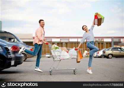 Cheerful family couple with bags in cart on supermarket car parking. Happy customers carrying purchases from the shopping center, vehicles on background. Cheerful family couple with cart on car parking