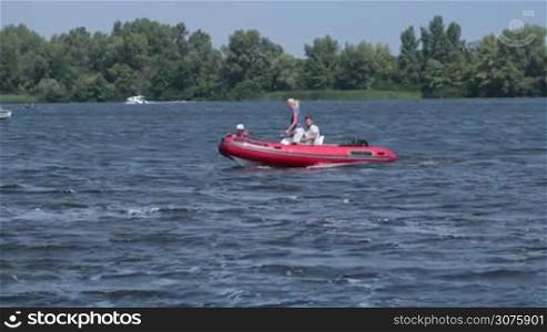 Cheerful family floating on red inflatable boat with motor in sunny day on a river during weekend