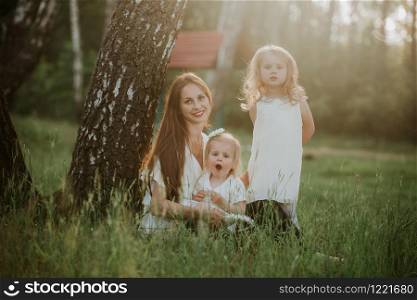 Cheerful family in a park, mom and their two lovely daughter to sit on the grass while they are looking at camera. happy family, mom and two daughters.. Cheerful family in a park, mom and their two lovely daughter to sit on the grass while they are looking at camera.