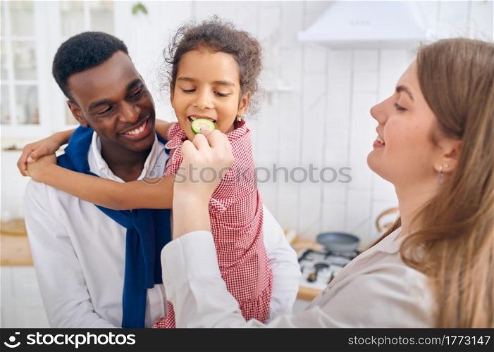 Cheerful family, nice breakfast on the kitchen. Mother, father and their funny daughter cooking in the morning, good relationship. Little child tastes a cucumber. Cheerful family, nice breakfast on the kitchen