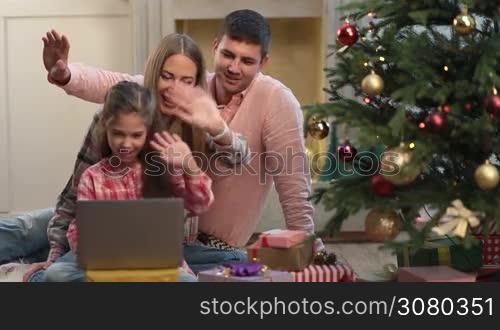 Cheerful family of three talking via skype with grandparents on Christmas eve. Smiling parents and adorable little daughter waving hello with hands and communicating over video call using laptop pc in xmas decorated room.