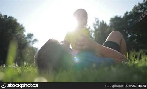 Cheerful farther and his little infant baby boy playing outdoors in glow of beautufil sunset while spending leisure together in summer park. Low view. Affectionate dad lying on back with smiling toddler son on chest, having fun and relaxing on lawn.