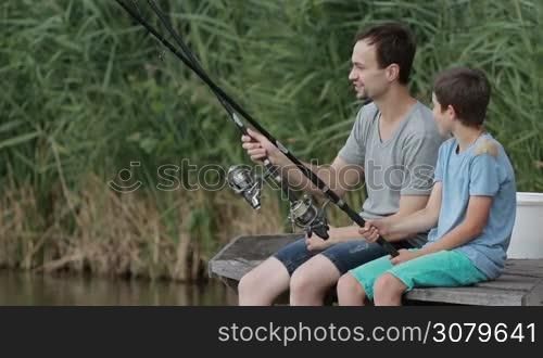 Cheerful father and his teenage boy talking while sitting on wooden pier with fishing rod at lake in summertime. Fishing by the lake is their common passion. Caring dad and son spending leisure together fishing in pond. Slow motion.