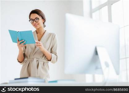 Cheerful female director holds personal organizer, looks through plans for day, wears beige formal costume, spectacles poses near dektop with big monitor white background verifies recorded information