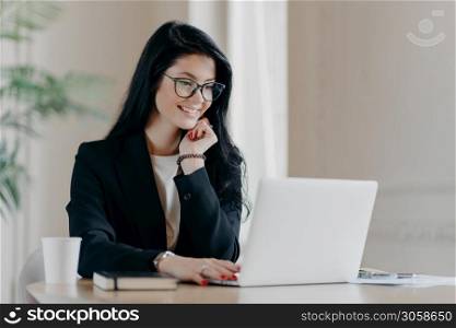 Cheerful female employee types on laptop computer, works remotely, concentrated with smile at device screen, writes email letter, works in office drinks coffee uses new application. Technology concept