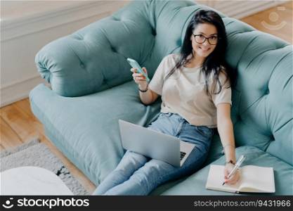 Cheerful female freelancer in casual outfit, writes in notepad, uses laptop, on couch. Student prepares project work at home.