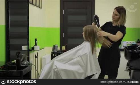 Cheerful female hairdresser drying cute girl&acute;s hair with blow dryer in barber shop. Side view. Professional hairstylist drying long straight blond hair with hair dryer in beauty salon over professional workplace background.