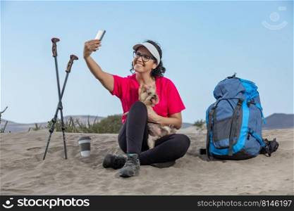 Cheerful female hiker sitting with backpack and trekking poles on sand on Lanzarote and taking self portrait with Yorkshire Terrier dog. Happy traveling woman taking selfie with dog